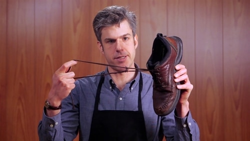 A white man holding a shoe in one hand and its laces stretched with the other hand. He is in front of a wood paneled wall and he's wearing an apron over a button down shirt.
