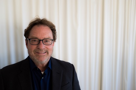 A white man in front of a white curtain. He is smiling and wearing black framed glasses