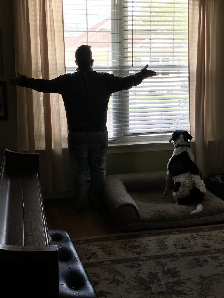 back view of a man looking out a window with his arms out, a dog next to him also looking out the window