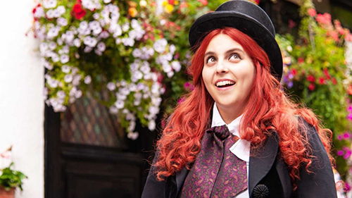 A still from 'How to Build a Girl' (2020). Beanie Feldstein with crazy red hair and a top hat.