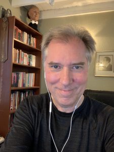 A selfie of John Cox in front of a bookshelf with a houdini prop of a human head in the background.