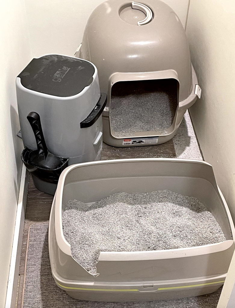 an open litter box in front of a litter box with a top on it, with a cat litter genie container to the left. all contained in a very narrow space
