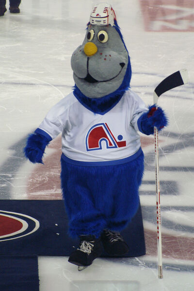 a large hockey mascot on the ice rink. the mascot is a blue otter with a gray face and yellow round nose, wearing a Nordiques jersey and hockey helmet, and holding a hockey stick