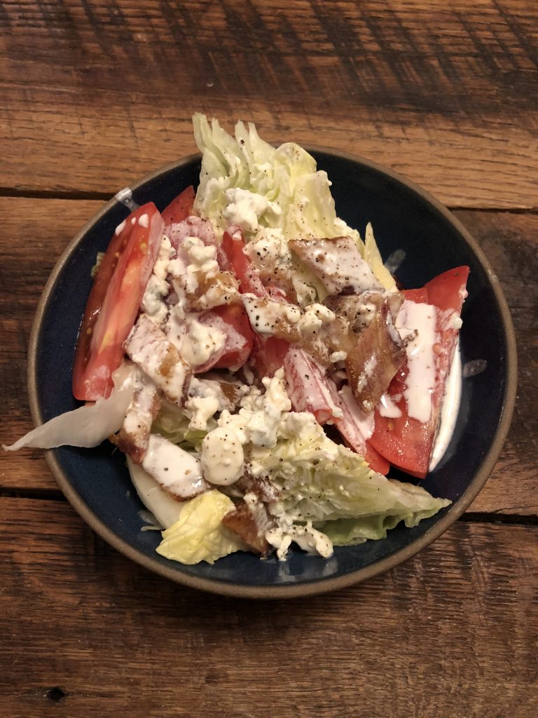 a wedge salad in a bowl sitting on a wooden table. In the bowl are a couple wedges of iceberg lettuce, topped with wedges of tomato, bacon pieces, and blue cheese dressing