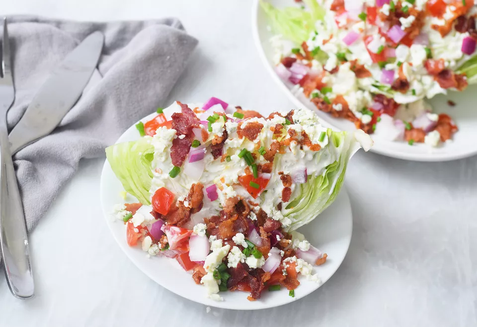 Two wedge salads on white plates, one in focus in the foreground: a wedge of iceberg lettuce topped with blue cheese dressing, bacon pieces, chopped tomato, chopped red onion, blue cheese crumbles, and chives