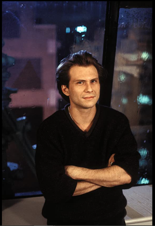 A white man (Christian Slater) with a 90s style floppy hairdo standing with his arms crossed and vaguely smirking.