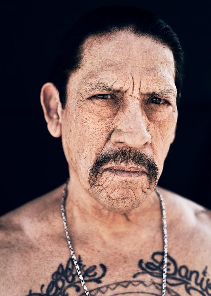 A Mexican American man with a handlebar mustache and dark hair pulled back in a ponytail. He has a serious expression on his face and is shirtless, wearing a chain necklace around his neck.