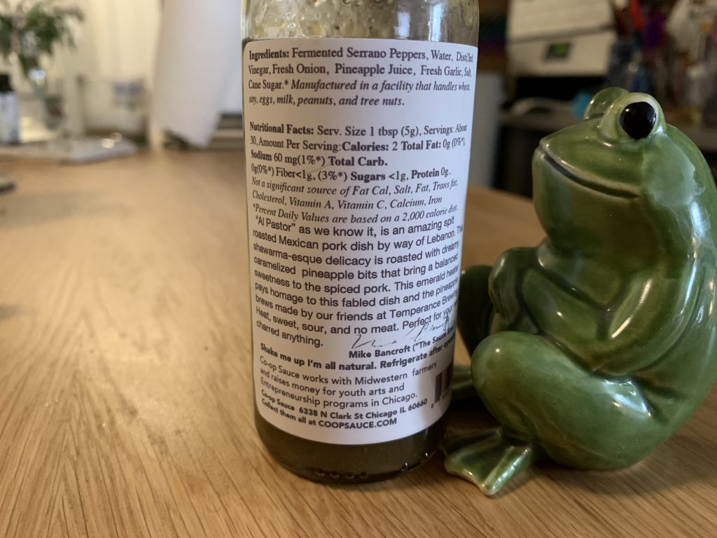 The back label of the hot sauce, listing the ingredients. To the right of the bottle is sitting a different ceramic frog with its legs folded in a criss cross applesauce style.