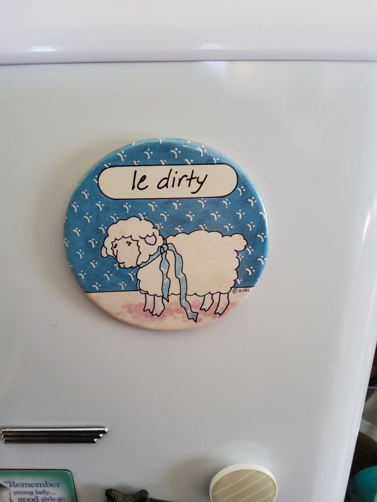 A round magnet on a white refrigerator. The magnet has a cartoon sheep on it and it says on top 