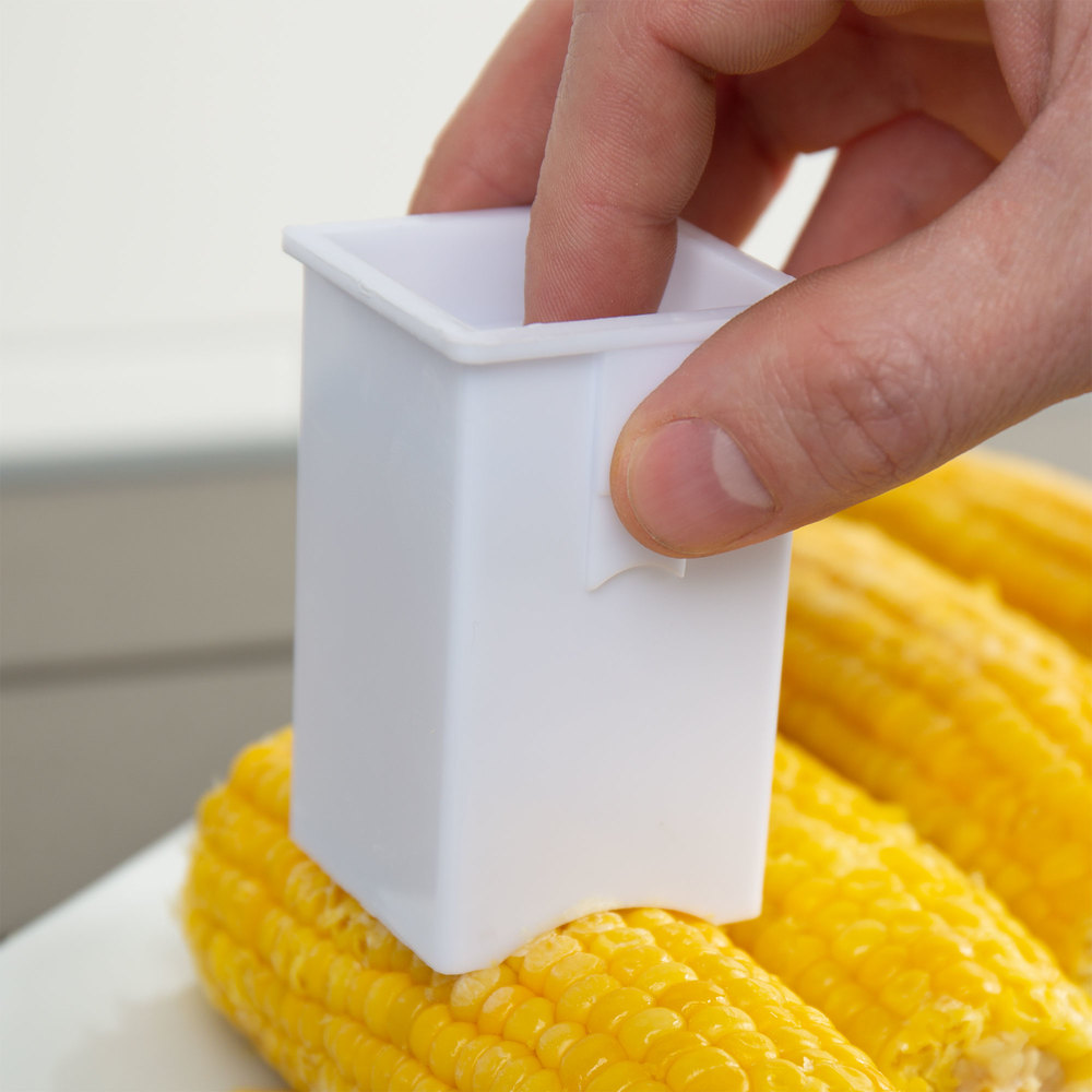 A hand holding a small white box over a piece of corn on the cob. The box is butter stick sized, with a curved bottom edge. This is the edge on the corn cob.