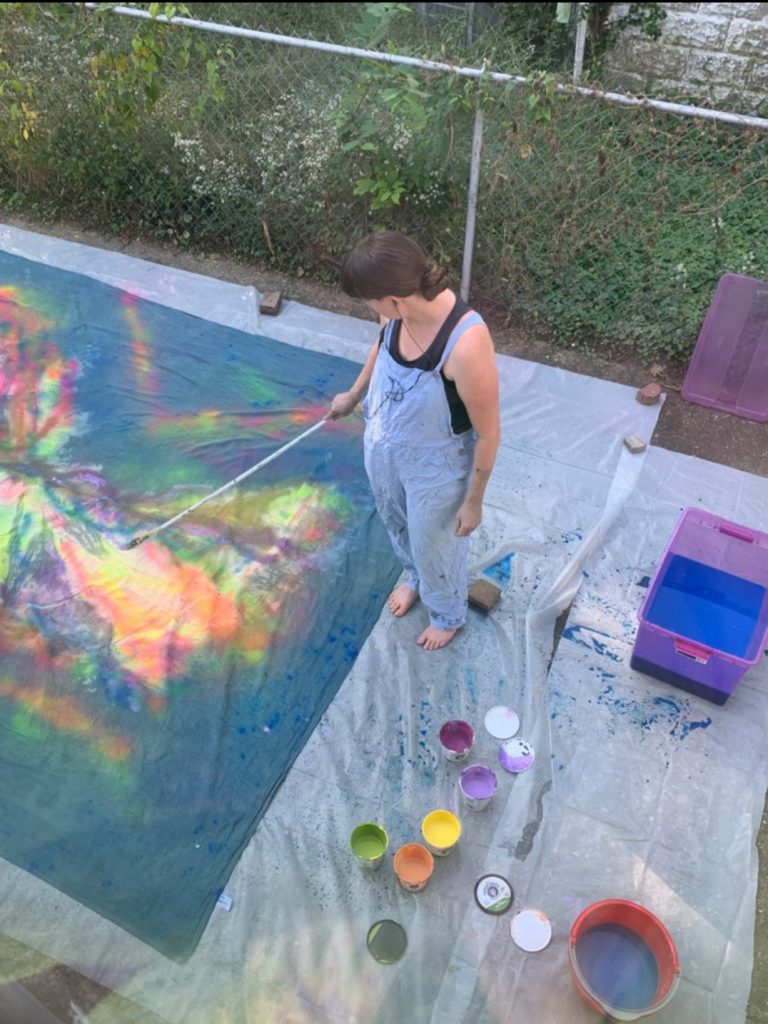 A woman with a long paintbrush on a stick standing outside over a large painted dropcloth, with paint containers all around her.