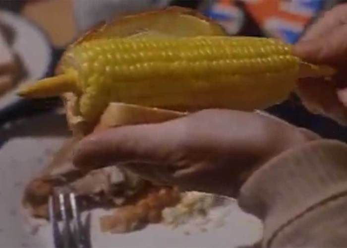 A hand holding a buttered slice of white bread around a piece of corn on the cob