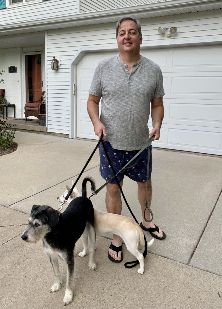 A man wearing a gray short and dark blue decorative sleep shorts with two leashed dogs