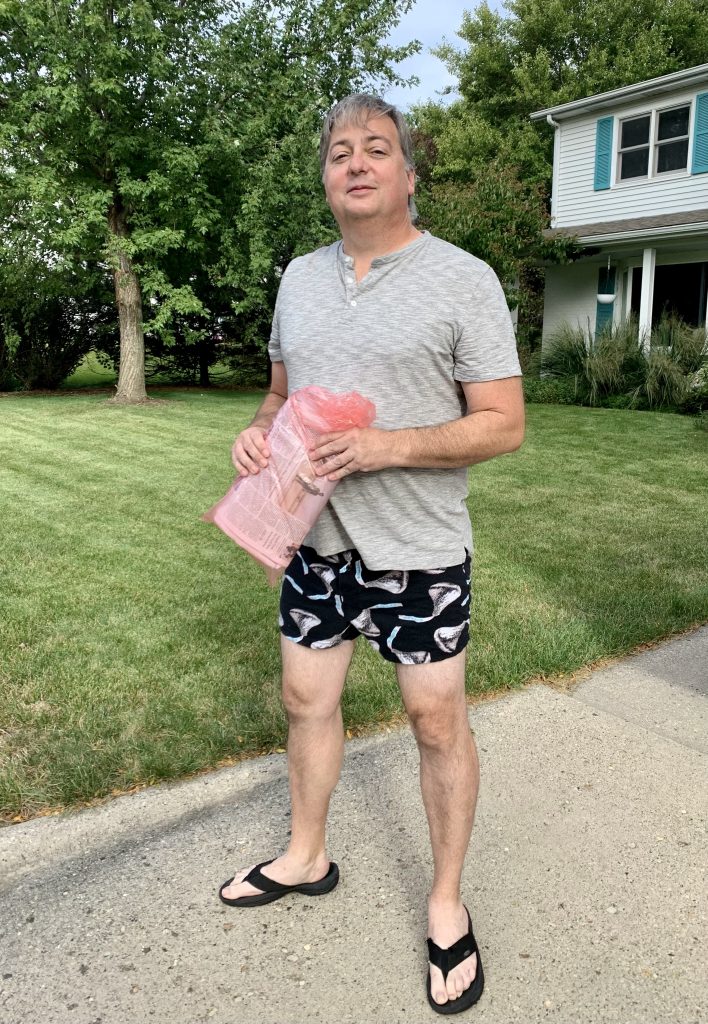 A man holding a still-wrapped newspaper outside his home, wearing a gray shirt and sleep shorts that are black in background with Hershey's Kisses as the design