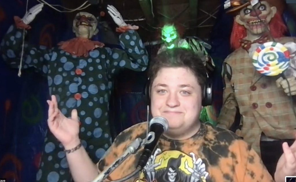 A teen boy wearing an orange and black tie dyed Spirit Halloween tshirt surrounded by three large animatronic scary clowns