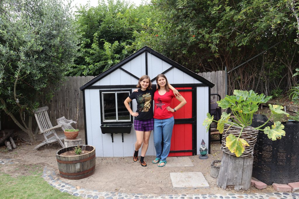 Two young women standing in front of a kids' playhouse turned shed that has been painted light grey with black trim and a red door