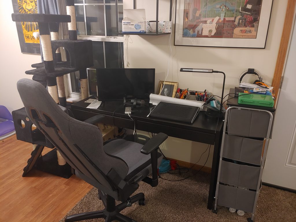 Another angle of the previous desk, showing to the left of it a large kitty condo. To the right of the desk are portable drawers. There is also framed wall art and a desk chair.