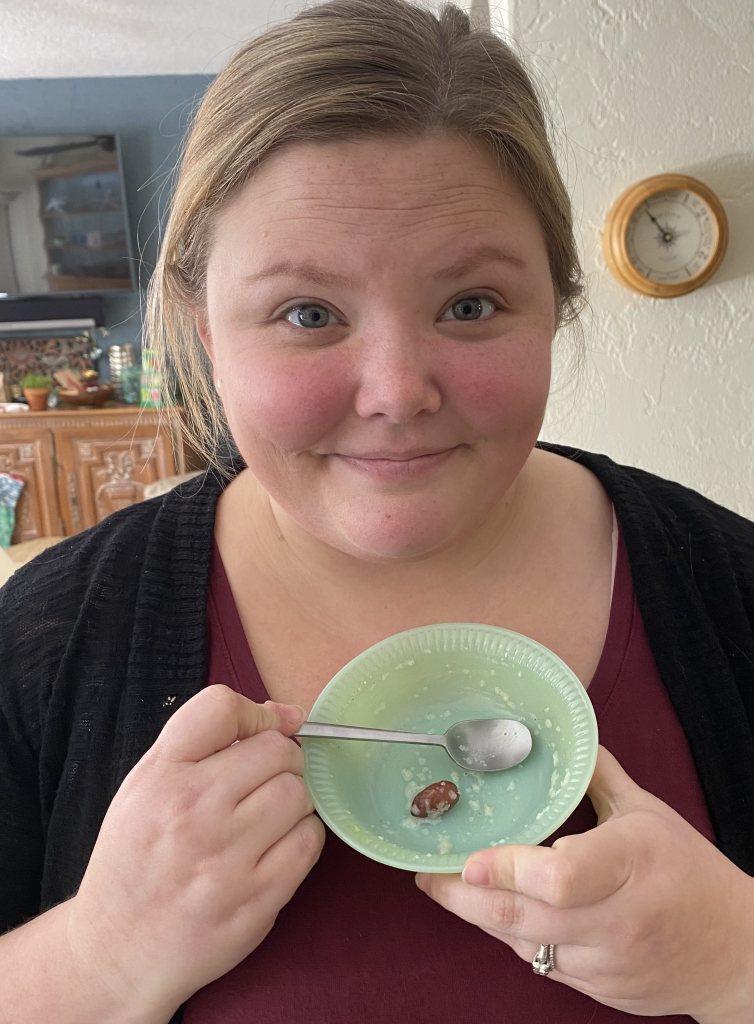 A blond woman holding a small, empty, green bowl with a spoon and a single almond inside the bowl.