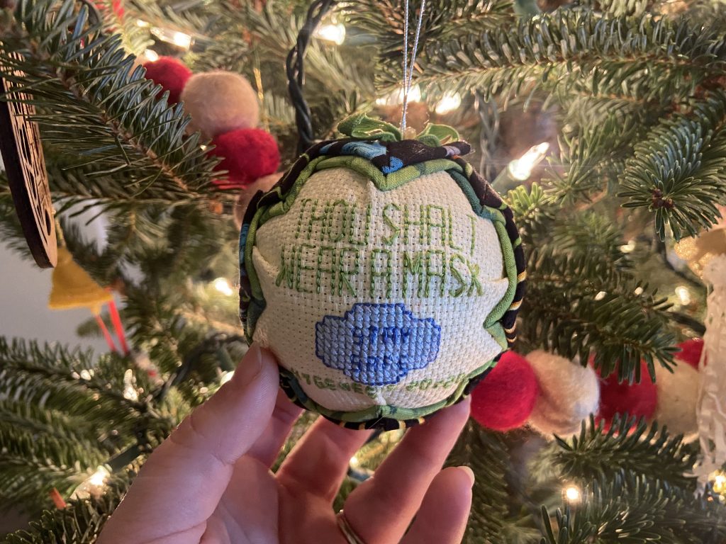 A cross stitched homemade ornament hanging from a lit Christmas Tree that says, 