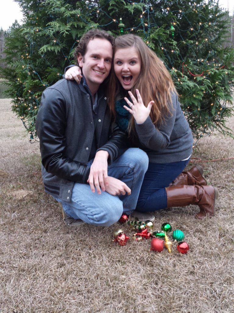 A man and woman crouched in front of a decorated tree outside with ornaments in front of them. The woman is holding her hand up in the classic 