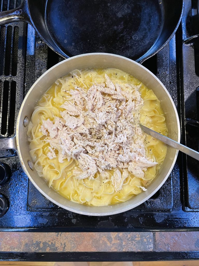 A pan on a stovetop with egg noodles and shredded chicken in it.