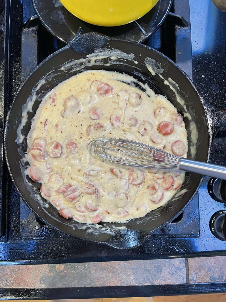 A white gravy on a stovetop with cut hot dogs in it.