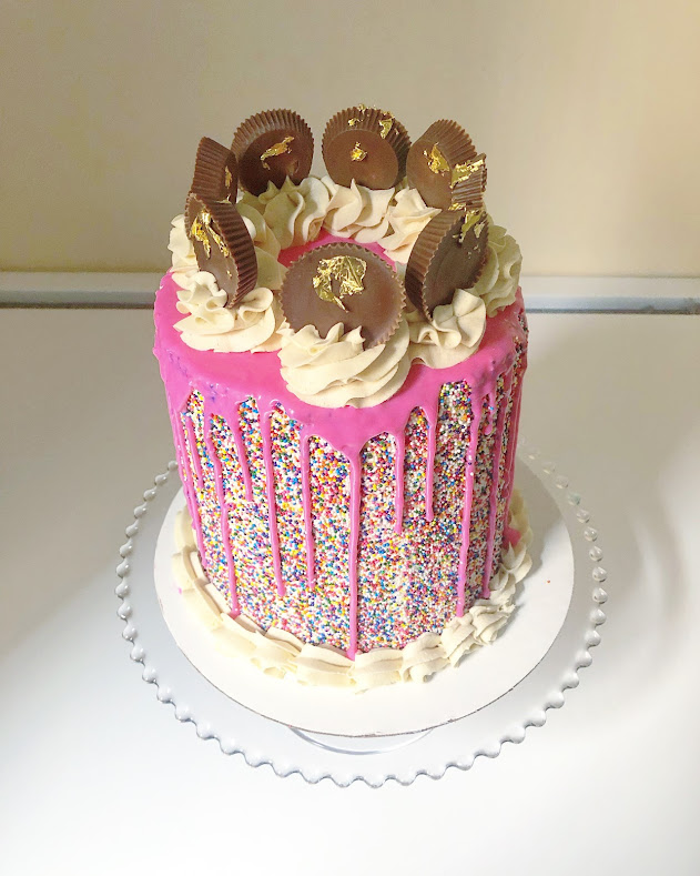 A layer cake covered in rainbow sprinkles, with hot pink icing on the top made to look like it's dripping down the sides. There are also on top peanut butter cups standing upright in white swirled icing