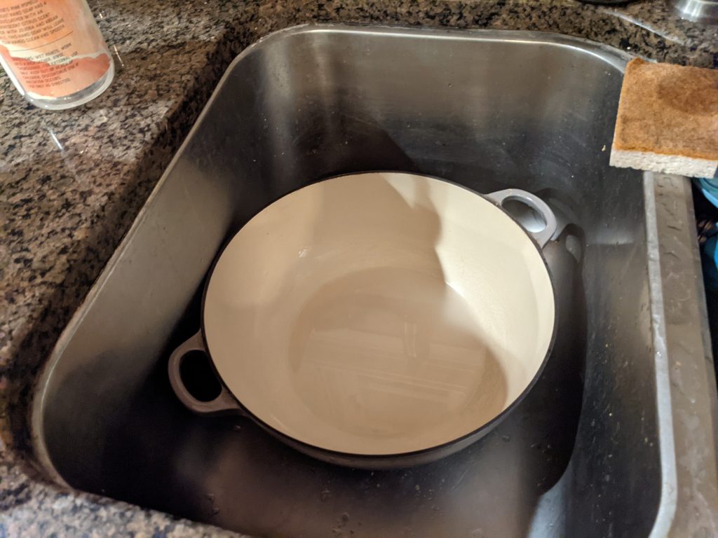 A clean looking Le Creuset pot sits in a deep stainless steel sink.
