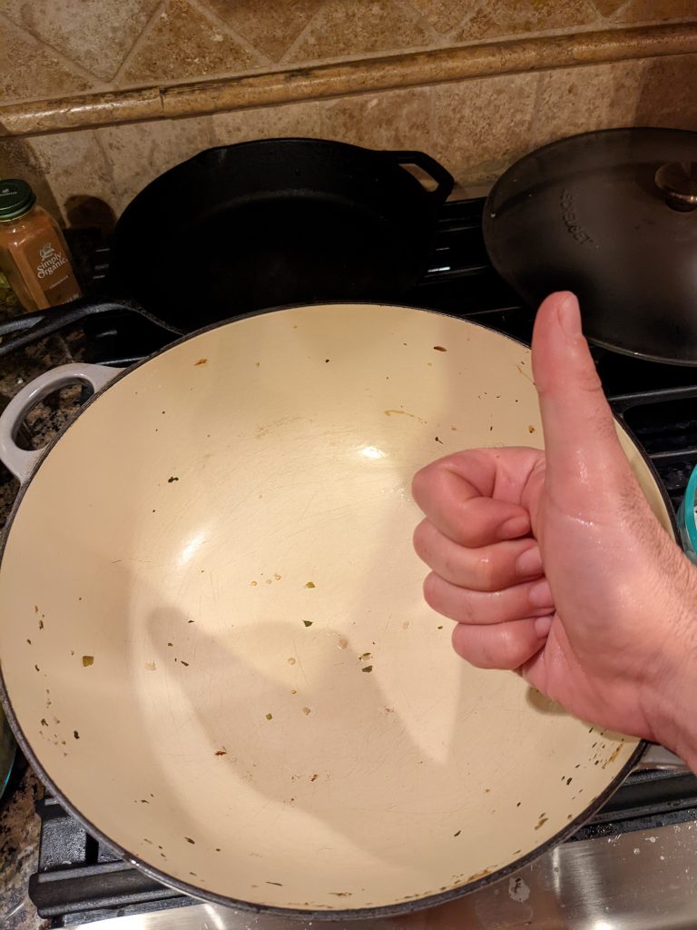 The same Le Creuset pot after it's been licked by the dog. It's is much cleaner, although there are small green remnants, and there is a person's hand doing a thumbs up sign in the foreground.