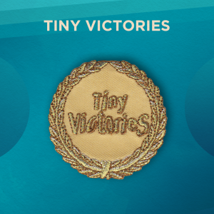 Tiny Victories. A crown of leaves with metallic silver, gold, and copper thread circles the border of this round patch. The center of the patch reads “Tiny Victories”. The background of the patch is gold.