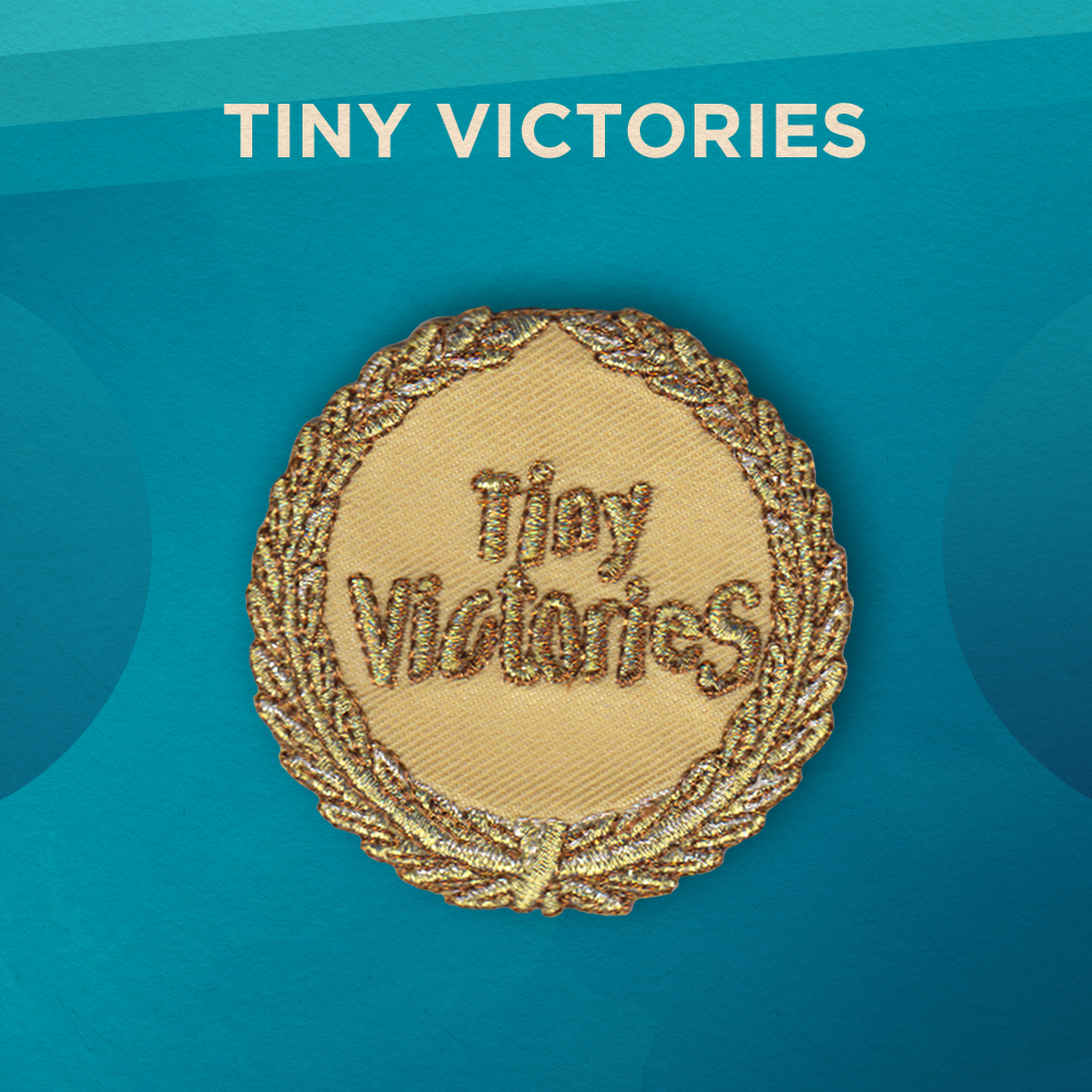 Tiny Victories. A crown of leaves with metallic silver, gold, and copper thread circles the border of this round patch. The center of the patch reads “Tiny Victories”. The background of the patch is gold.