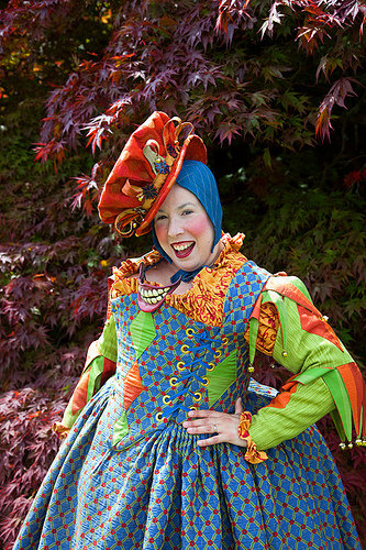 A woman in a bright blue Renaissance Court Jester costume in front of a bunch of trees with purple leaves