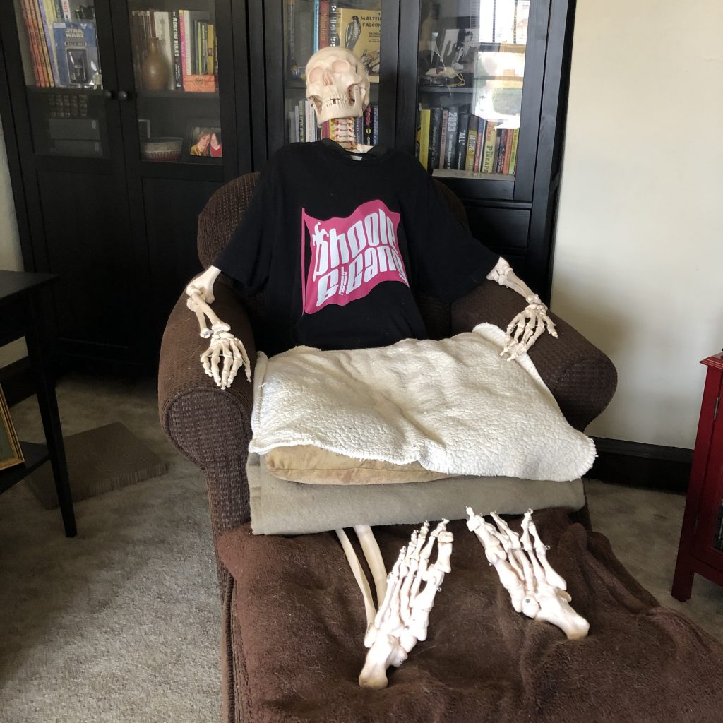 A skeleton wearing a t-shirt placed in a sitting position on an armchair, with its feet up on an ottoman and a pillow in its lap
