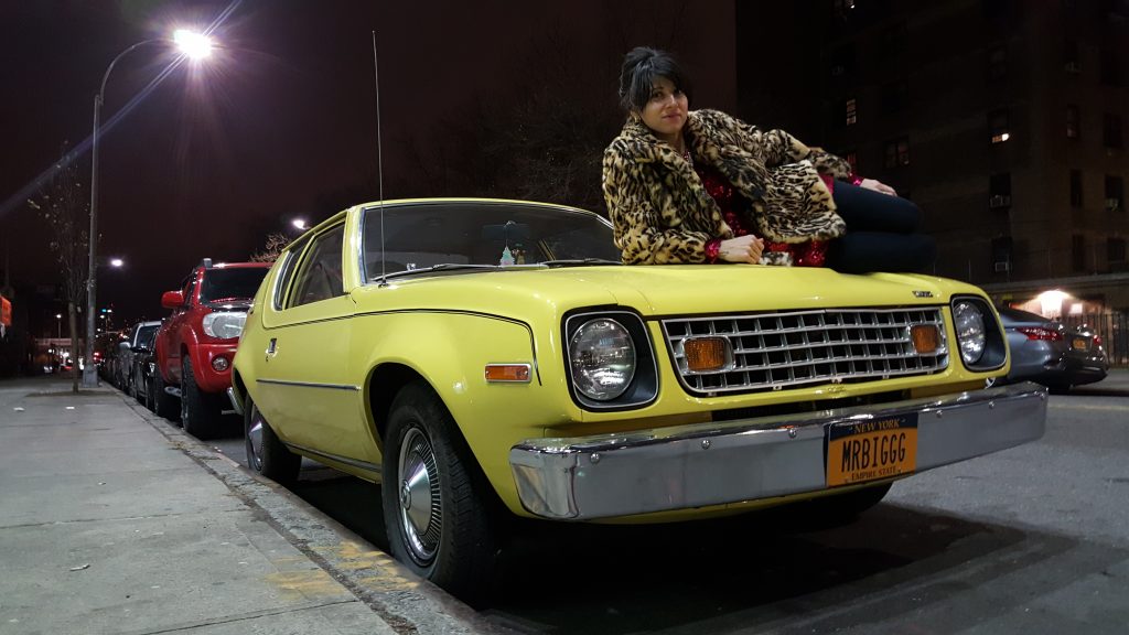 A woman posing for a photo on the hood of a yellow Gremlin with the license plate 