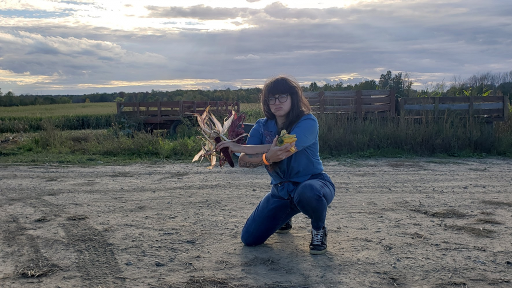 A woman crouching with farmland in the background, holding a corn cob, looking cool