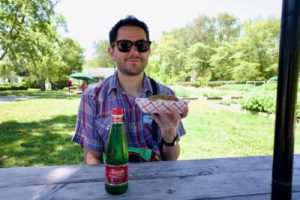 Abe Forman-Greenwald as an adult. He's at a park holding a hot dog. He's seated at a picnic table with a seltzer water in front of him and wearling a plaid purple and red shirt.