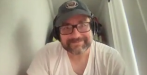 Alex Goldman in his closet wearing a hat, glasses and headphones. He is in a white shirt.