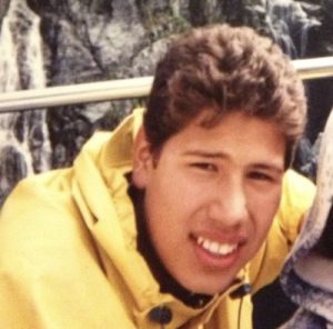 Migizi as a teenager. He is in a yellow jacket by what appears to be a cliffside. 