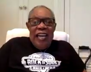 Sam Moore wearing a black shirt. He's smiling and sitting on a white chair.