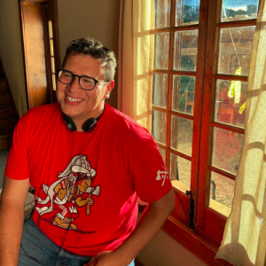 Migizi in a red shirt sitting by a window. He has headphones around his neck and is smiling. He is wearing glasses.