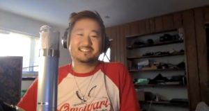 Kishi Bashi is smiling and looking straight at the camer. This is a screenshot from a zoom window. He's wearing a red raglan shirt with a white middle. 