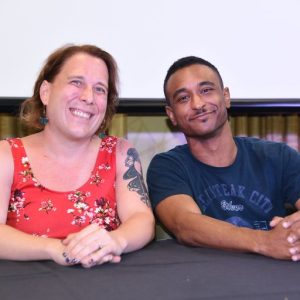Amy Schneider and Brandon Blackwell. They're both sitting at a table smiling at the camera. Amy is wearing a lovey floral dress and Brandon is wearing a cooll T Shirt