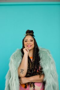 Ashley Ray as an adult. Shes in front of a blue background and wearing a fur coat. Even more fabulous hair than when she was a teenager.