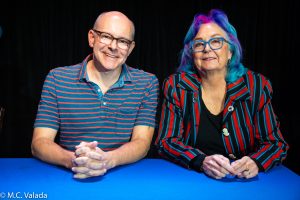 Robb Corddry and Mimi Pond. Rob is bald and wearing a colored collared shirt. Mimi has a lovely colored hair of purple an blue and is wearing a striped color shirt.