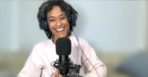 Abbi Crutchfield on the zoom window. She's smiling at the camera and is in front of a microphone. She's wearing headphones over her curly hair.