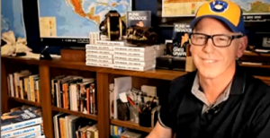 Greg Graffin in his office. He's smiling at the camera and is wearing an old-school Brewers cap. His shelf with his many books is in behind him.