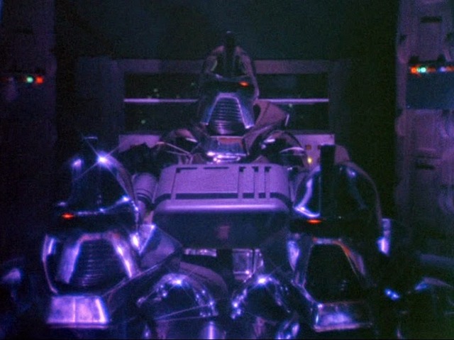 A screenshot of three Cylons, from the TV show Battlestar Galactica, piloting a Cylon Raider spaceship. Two Cylons are sitting side-by-side in the foreground, and the third is sitting behind them, in the center of the frame. The scene is mostly in shadow, with violet light reflecting off the Cylons' metallic bodies.