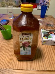 An old, mostly full bottle of honey with a yellow cap, on a kitchen table. The honey is Kirkland Signature brand and is separated into three crystalized layers.