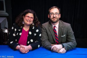 Muffy Marracco & Brad Rutter. Muffy is wearing a polka-dot cardigan and red glasses. Brad is wearing black glasses and a suit with a red and blue striped tie. They are both sitting at a table with a blue tablecloth