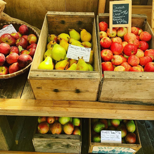 A photo of two wooden shelves, part of a larger farm stand. The top shelf has a basket of red apples on the left, a wooden box of Bartlett pears in the middle, and a wooden box of sunrise apples on the right. The bottom shelf has a wooden box of red-and-green apples on the left, and a wooden box of Bennett pears on the right.